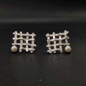 Può includere: Un paio di orecchini d'argento con un design a griglia quadrata e un singolo perla che pende da ogni orecchino.