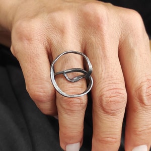 Op de afbeelding: Een zilveren ring met een uniek, abstract ontwerp. De ring is gemaakt van één stuk metaal dat is gedraaid en gebogen tot een cirkel.