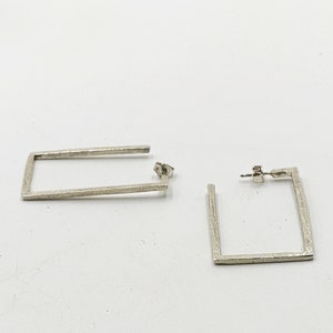 May include: A pair of silver square hoop earrings. The earrings are made of a textured metal and have a simple, modern design.