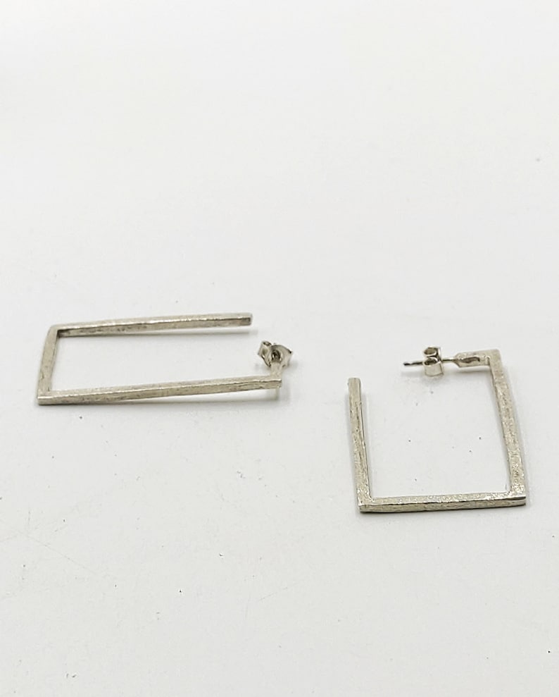 May include: A pair of silver square hoop earrings. The earrings are made of a textured metal and have a simple, modern design.