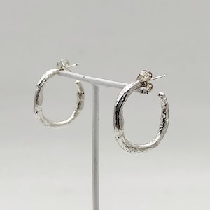 May include: A pair of silver hoop earrings with a textured finish, displayed on a white stand. The earrings have an organic, irregular shape. The background is plain white, highlighting the earrings' design. The earrings are made of silver.