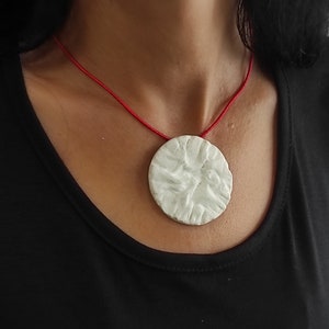 May include: A white, circular pendant with a textured surface hangs from a thin, red cord. The pendant is centered against a black top, creating a striking contrast. The necklace is a simple yet elegant piece of jewelry.