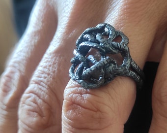 May include: A silver ring with a textured, intricate design. The ring is worn on a finger.
