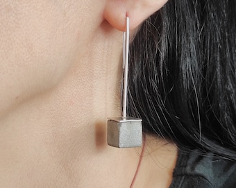 May include: A pair of silver earrings with a small, gray concrete cube hanging from each. The earrings are dangling from the earlobes of a person with long black hair.