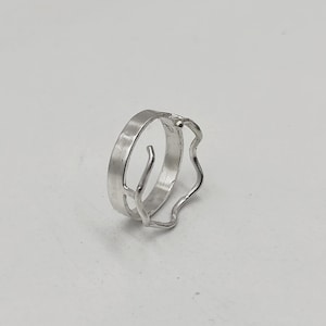 Puede incluir: Un anillo de plata con un diseño único. El anillo presenta una banda lisa y plana y un borde ondulado y texturizado. Un pequeño trozo de metal curvado se extiende desde la banda, agregando un elemento artístico. El anillo se muestra sobre una superficie blanca.