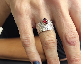 May include: A silver ring with a textured band and a red gemstone. The ring is worn on a finger with dark blue nail polish.