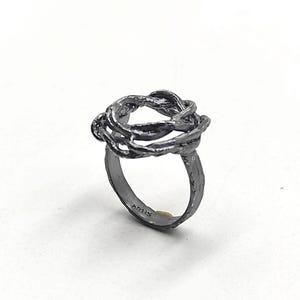 Op de afbeelding: Een donkergrijze ring met een getextureerde band en een geweven, knoopachtig ontwerp aan de bovenkant. Het woord "ARTIS" is op de band gegraveerd. De ring staat tegen een witte achtergrond.