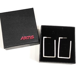 May include: A pair of silver rectangular hoop earrings in a black box with the word "ARTIS" printed on the top.