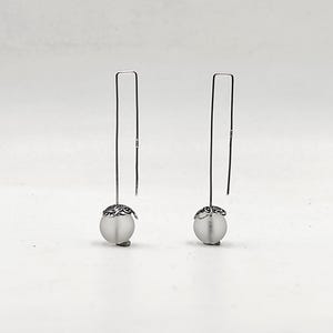Könnte beinhalten: Ein Paar silberne Ohrringe mit einer mattierten weißen Perle und einem zarten silbernen Design an der Oberseite jeder Perle.