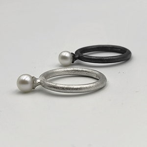Könnte beinhalten: Zwei schlichte Silberringe mit je einer Perle. Der Ring links hat eine gebürstete Silberoberfläche, während der Ring rechts eine schwarz oxidierte Oberfläche hat.