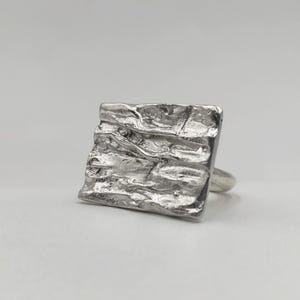 Könnte beinhalten: Ein silberner Ring mit einer strukturierten, quadratischen Oberfläche. Die Oberfläche hat eine raue, unebene Textur, die ihm ein einzigartiges, künstlerisches Aussehen verleiht. Der Ring hat ein glattes, schlichtes Band.