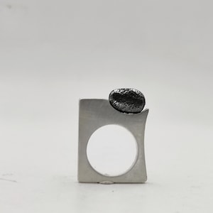 Può includere: Un anello moderno in argento con una fascia rettangolare e una grande pietra scura e strutturata sulla parte superiore. L'anello presenta un'apertura circolare al centro della fascia. L'anello è posto su uno sfondo bianco.