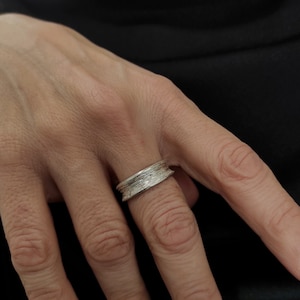 Op de afbeelding: Een zilveren ring met een getextureerd, gelaagd ontwerp. De ring wordt aan een vinger gedragen.