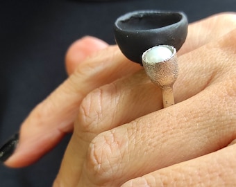 May include: A black ceramic ring with a silver band and a white pearl accent. The ring is worn on a person's finger.
