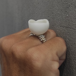 Op de afbeelding: Een witte keramische ring met een unieke, organische vorm. De ring is gezet op een zilveren band met een textuurontwerp.