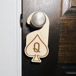 May include: A wooden door hanger shaped like a spade with a gold 'Q' in the center. The hanger is attached to a silver door knob.