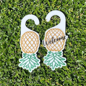 Puede incluir: Dos colgadores de puerta blancos con diseño de piña. La piña tiene un cuerpo dorado con purpurina y hojas verdes con purpurina. Un colgador tiene la palabra "Welcome" en escritura negra.