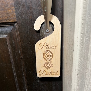 May include: A wooden door hanger with the text "Please Disturb" and a pineapple graphic. The hanger is brown and has a hole at the top for hanging on a door knob.