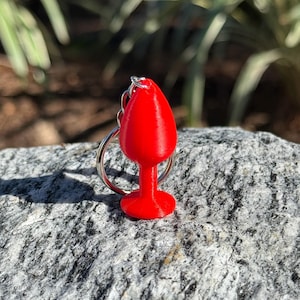 May include: A bright red, 3D-printed keychain charm shaped like a miniature wine glass. The charm is attached to a silver-colored keyring. The background is blurred, with green foliage and sunlight.