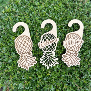 May include: Three wooden pineapple door hangers with a welcome sign. The hangers are painted in a light wood color and have a brown pineapple design with the word "Welcome" written on each.