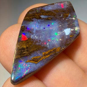 May include: A polished boulder opal gemstone with a kite-like shape, featuring a dark brown ironstone matrix interspersed with vibrant flashes of blue, green, and red play-of-color across its smooth, reflective, and slightly irregular surface.