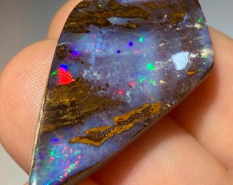 May include: A polished boulder opal gemstone with a kite-like shape, featuring a dark brown ironstone matrix interspersed with vibrant flashes of blue, green, and red play-of-color across its smooth, reflective, and slightly irregular surface.