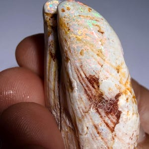 Puede incluir: Un espécimen de ópalo en bruto y alargado con una base de color blanco crema y destellos iridiscentes en verde, naranja y rojo. La superficie muestra inclusiones naturales y texturizadas de hierro marrón y vetas por toda la forma irregular.