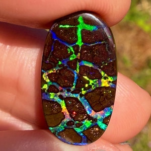 Może przedstawiać: Owalny kamień szlachetny opal boulder z ciemnobrązową matrycą żelazistą, z żywymi, opalizującymi żyłkami w kolorach neonowej zieleni, elektrycznego błękitu i żółci, odbijającymi się na wypolerowanej, gładkiej powierzchni kamienia.