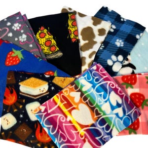 Puede incluir: Una colección de cuadrados de tela polar con varios diseños. Los diseños incluyen rebanadas de pizza, fresas, pandas, manchas de vaca, corazones y s'mores. Las telas son en tonos de azul, negro, rosa, blanco y marrón.