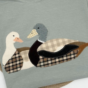May include: A pale green Comfort Colors t-shirt with an appliqué design of two ducks. The ducks have patterned details in black, brown, and beige. The shirt has a crew neck and a tag with the Comfort Colors logo.