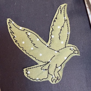 May include: An olive green fabric applique in the shape of a bird in flight, featuring small white polka dots and black machine-stitched outlines. The bird is positioned on a solid black fabric background held within a plastic embroidery hoop.