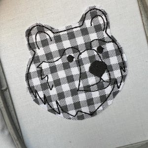May include: A fabric patch in the shape of a bear's head. The bear's face is a black and white checkered pattern, with black embroidered details for the eyes, nose, and outline. The patch is set in an embroidery hoop.
