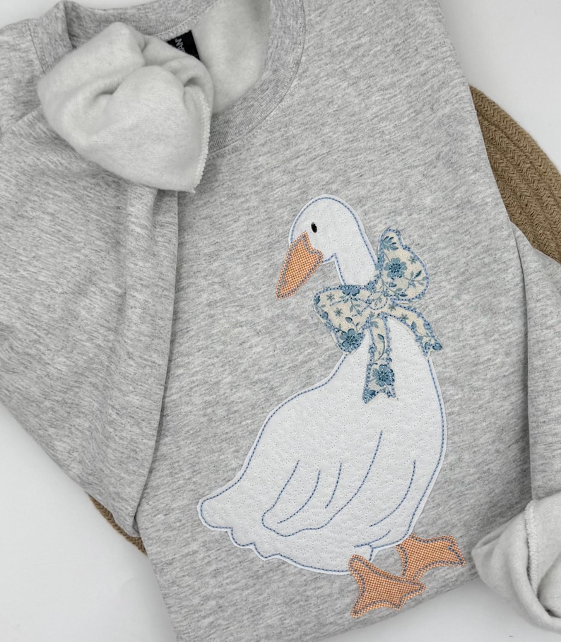 May include: A light gray sweatshirt featuring a white goose applique with a blue floral bow. The goose has an orange beak and feet. The sweatshirt has a crew neck and long sleeves.