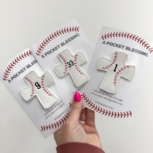 Puede incluir: Tres bendiciones de bolsillo en forma de cruz blanca con costuras de béisbol y los números 1, 9 y 33. Cada tarjeta tiene el texto "A Pocket Blessing" y un versículo bíblico. Las tarjetas son sostenidas por una persona con esmalte de uñas rosa.