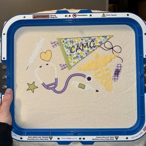 May include: An embroidered design featuring medical symbols like a stethoscope, caduceus, thermometer, and band-aid. A pennant with "CAMC" is also included. The design is set within a blue embroidery hoop.