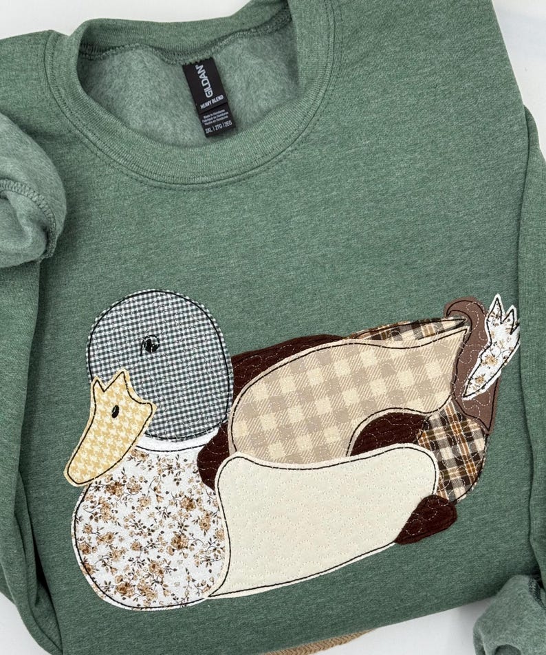 K&ouml;nnte beinhalten: Gr&uuml;nes Sweatshirt mit einem Patchwork-Enten-Design. Die Ente hat einen karierten Kopf, einen beige-braunen K&ouml;rper und eine Brust mit floralem Muster. Das Sweatshirt hat einen Rundhalsausschnitt und lange &Auml;rmel. Das Etikett lautet "Gildan Heavy Blend".