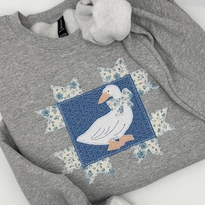 May include: A grey sweatshirt with a white goose design, complete with a blue bow and peach feet, set on a blue square. The square is surrounded by floral starbursts in blue and cream. The sweatshirt has a crew neck.