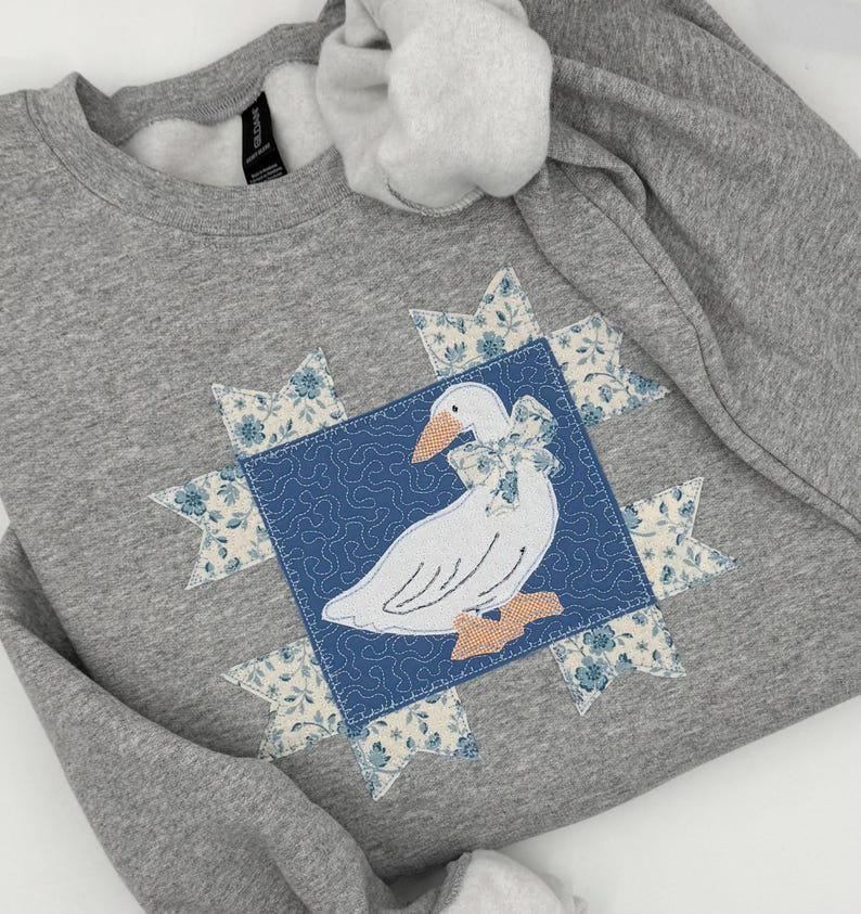 May include: A gray sweatshirt featuring a white goose applique with a blue bow and peach feet, centered on a blue square. The square is surrounded by floral starbursts in blue and cream. The sweatshirt has a crew neck.