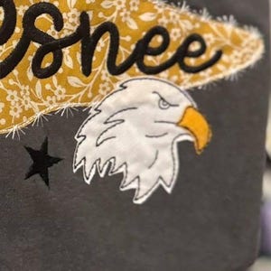May include: A dark gray fabric item featuring an embroidered white eagle head with a yellow beak, a black star, and a mustard yellow floral patterned fabric patch with the black embroidered text "esnee" stitched across the top section.