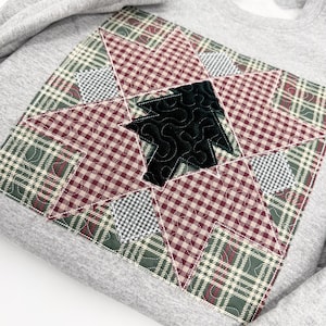 May include: A light gray sweatshirt featuring a large, quilted star applique on the front. The star is constructed from patches of red and white gingham, green and white plaid, and black and white checkered fabric, with a black velvet tree.