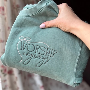 May include: A sage green sweatshirt with the embroidered words "WORSHIP anyways" in a script font. The sweatshirt is held in a hand with pink nail polish. The design features a bow above the word "WORSHIP".