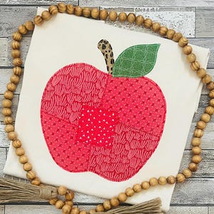 May include: A cream-colored t-shirt featuring a large apple applique made from various red patterned fabrics. The apple has a brown leopard print stem and a green quilted leaf. A string of wooden beads with tassels frames the shirt.