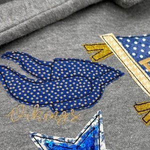 May include: Close-up of a gray sweatshirt with embroidered patches. The patches include a blue and white polka-dot design, a blue sequined star, and a flag with gold accents. The word "Vikings" is embroidered in gold.