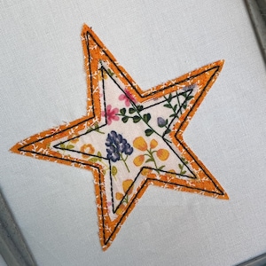May include: A fabric star applique with an orange border and floral pattern. The star is stitched onto a white background. The inner star features a colorful floral design with pink, blue, and yellow flowers.