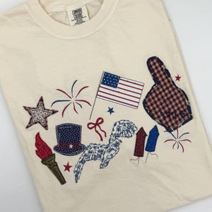 May include: Cream-coloured t-shirt with patriotic appliqués. Features an American flag, fireworks, a star, a top hat, a torch, and a foam finger. The shirt has the words "Comfort Colors" on the tag.