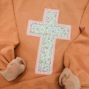 May include: A peach-colored crewneck sweatshirt featuring a large, centered cross appliqué. The cross is crafted from a light green fabric with a delicate floral pattern, bordered by a thin pink and white gingham check fabric trim.