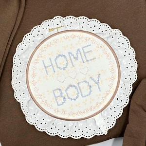 May include: A round embroidery hoop with the words "HOME BODY" stitched in blue thread. The design includes three small hearts and floral accents. The hoop is surrounded by a white, scalloped lace trim, creating a decorative piece.