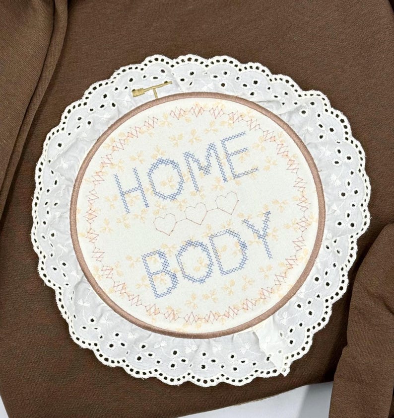 May include: A round embroidery hoop with the words "HOME BODY" stitched in blue thread. The design includes three small hearts and floral accents. The hoop is surrounded by a white, scalloped lace trim, creating a decorative piece.