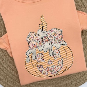 May include: A peach-coloured t-shirt featuring a central appliqué of a pumpkin topped with a melting candle. The pumpkin and candle are decorated with a floral pattern and white diagonal stripes, accented by a large floral bow at the top.