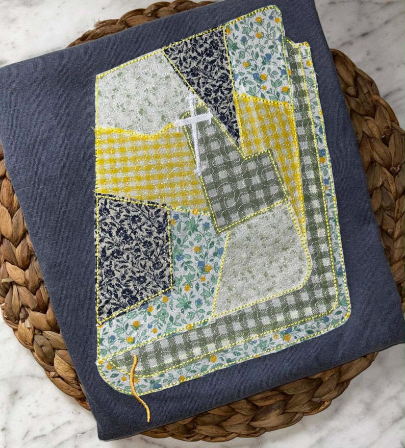 May include: A dark gray sweatshirt with a patchwork design featuring various fabric patterns. The patchwork includes yellow gingham, floral, and navy blue patterned fabrics. A white cross is embroidered on the patchwork. The edges are outlined with yellow stitching.